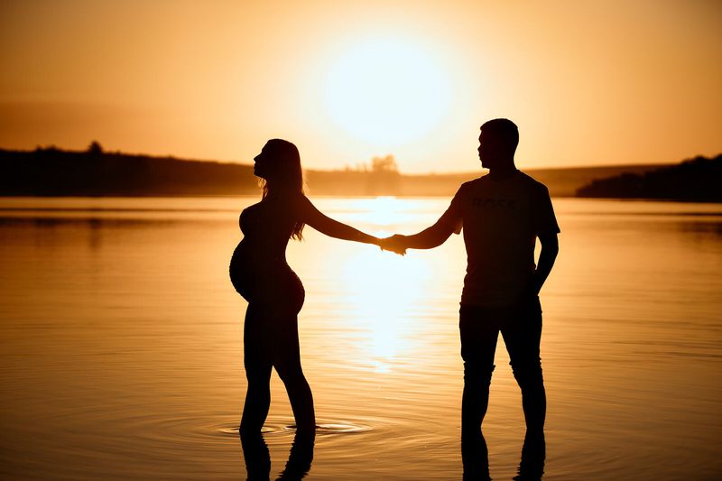 Silhueta de um casal de mãos dadas ao pôr do sol, ela grávida, com o reflexo do sol na água