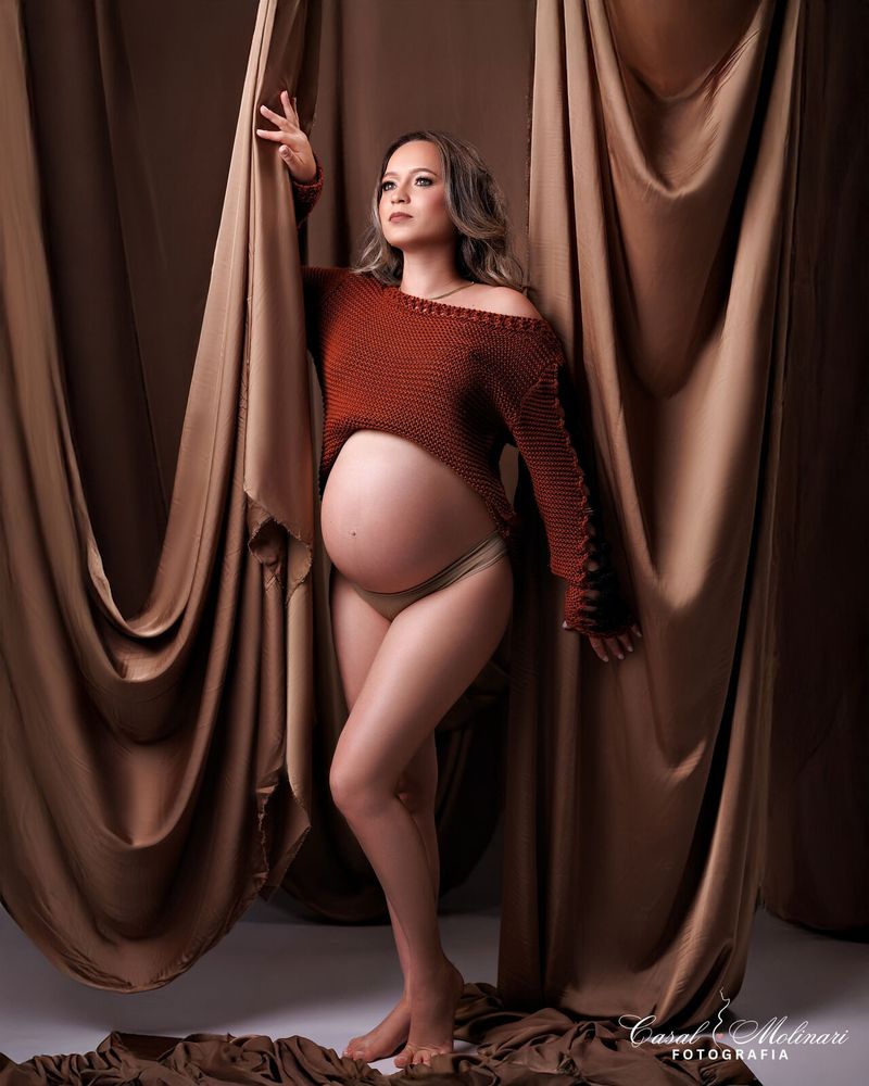 Gestante diante de cortinas marrons, com blusa de tricô e a barriga à mostra