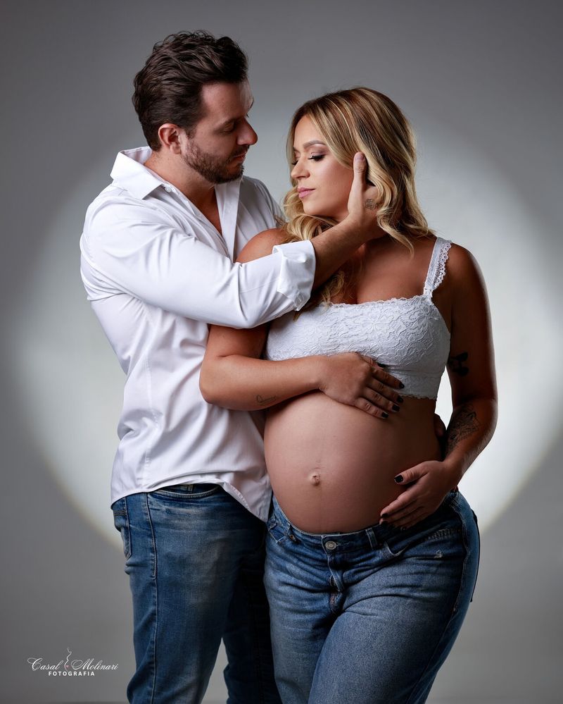Casal em estúdio, ele de camisa branca abraçando a gestante