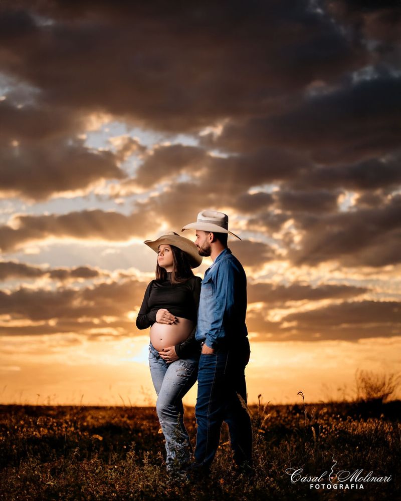 Casal de gestante com chapéu de cowboy em campo aberto ao pôr do sol
