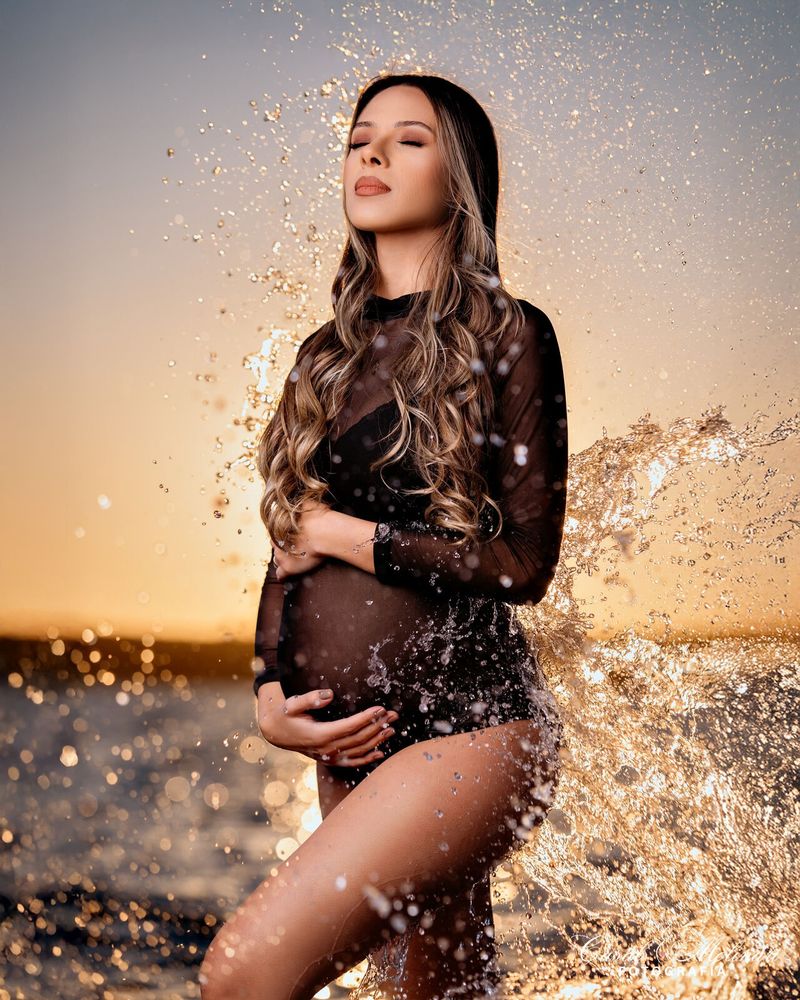 Gestante em ensaio splash, com água respingando ao pôr do sol