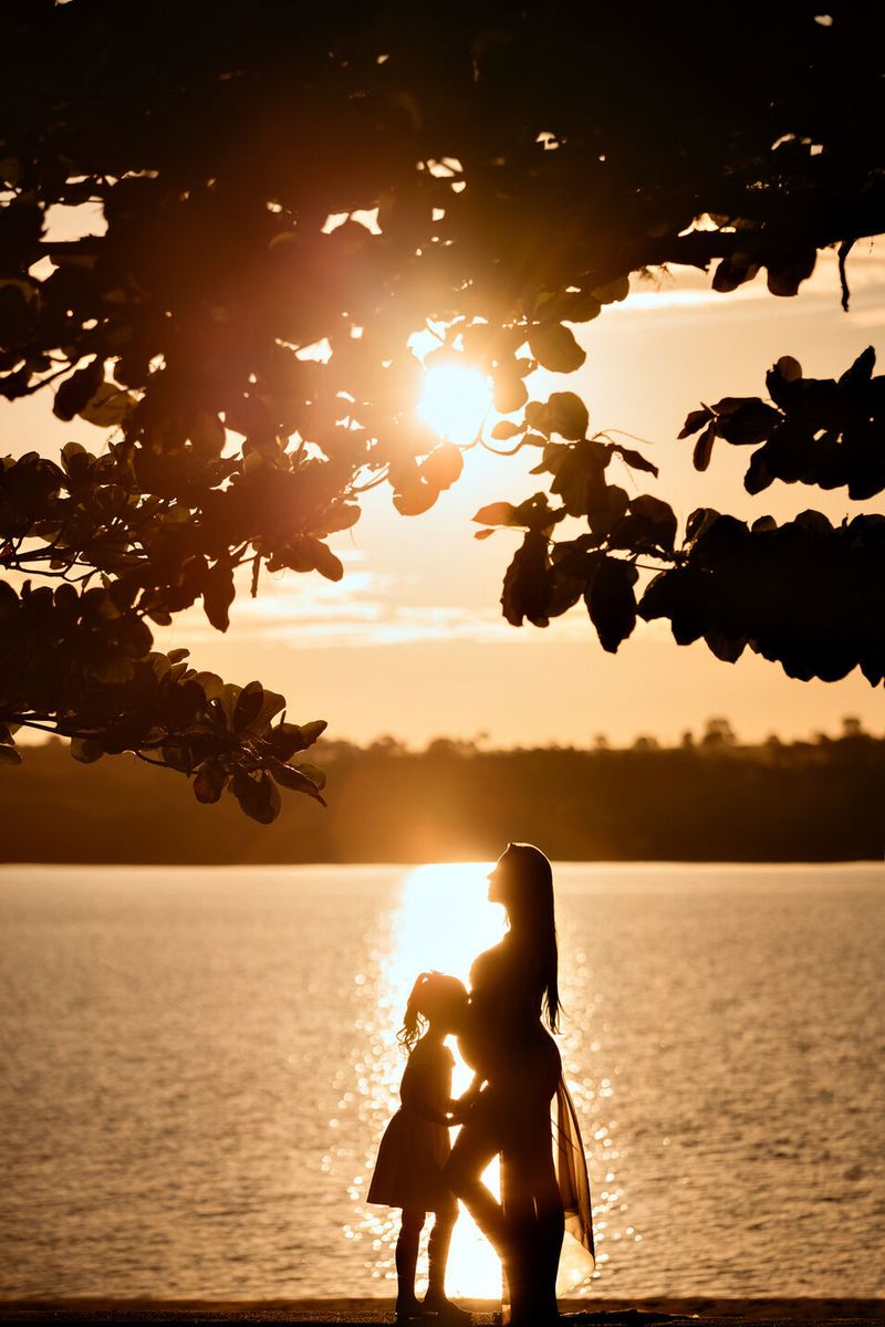 Silhueta de gestante com a filha à beira do lago ao pôr do sol