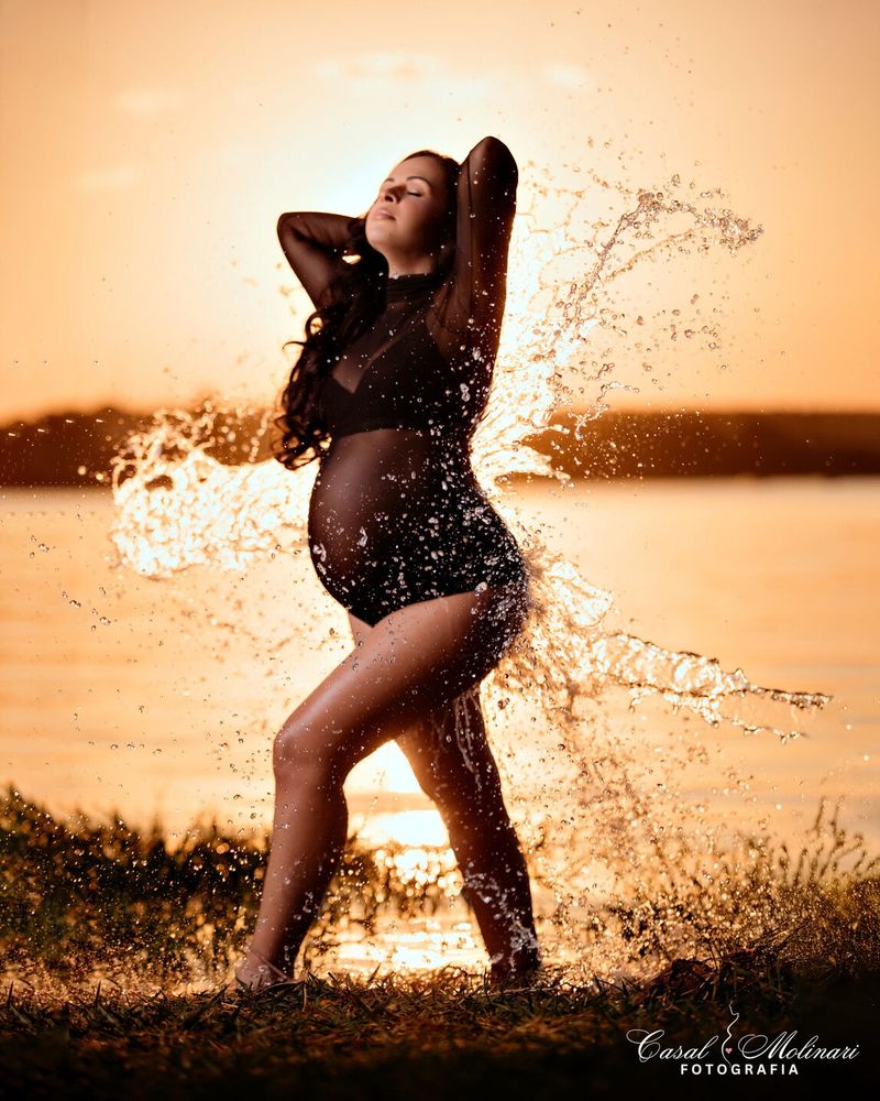 Gestante em ensaio splash, com água respingando ao pôr do sol